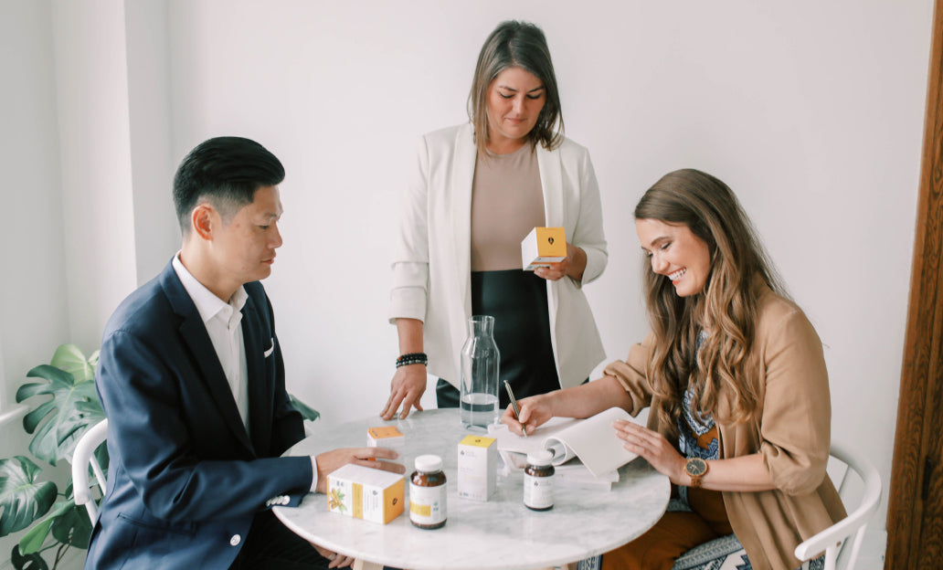 A man and two women sit around a table full of Living Alchemy Products reading the benefits and smiling.